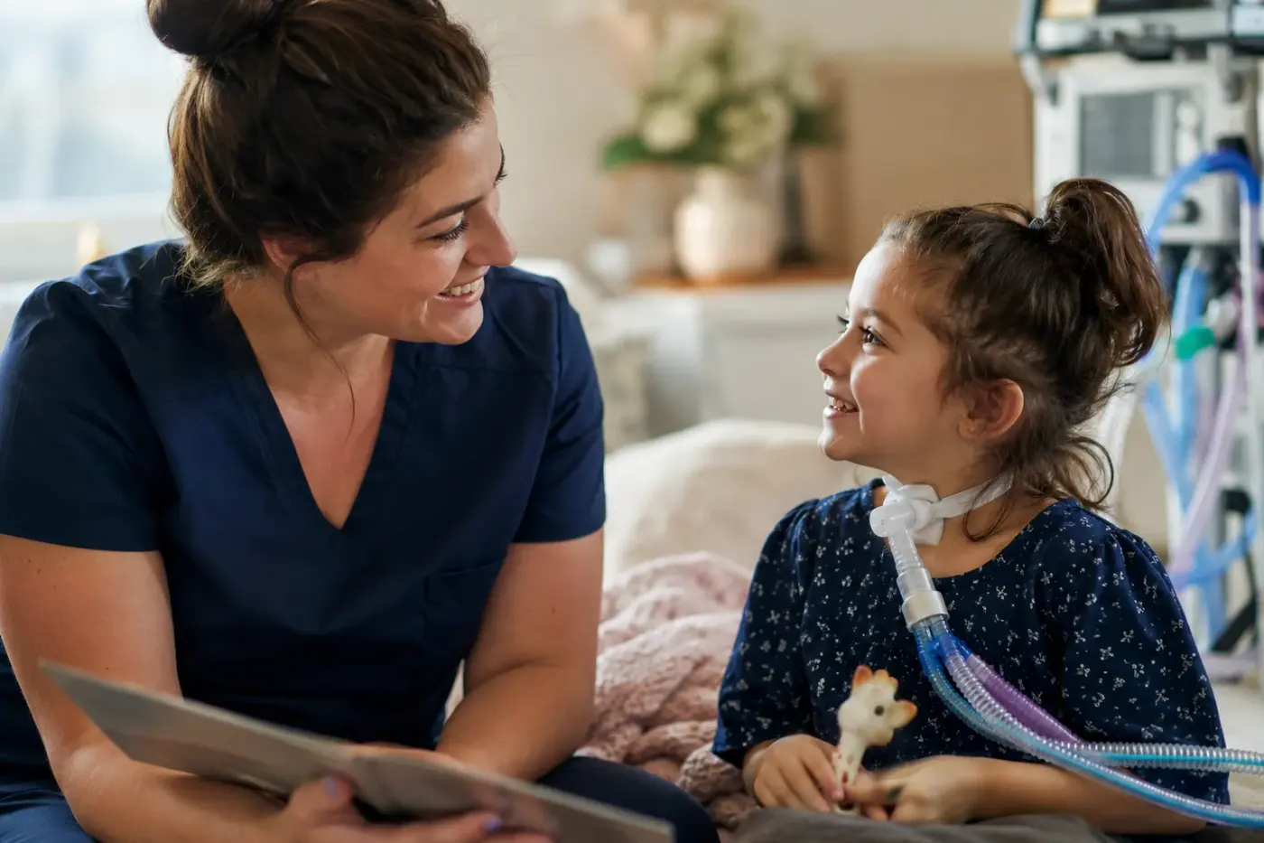 A nurse in navy scrubs reads a picture book with a young girl who has a tracheostomy and ventilator tubing.